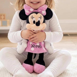 May include: A Minnie Mouse plush doll with a pink polka dot dress and bow. The doll has black ears, a beige face, and the name "Norah" embroidered on the dress. The doll is being held by a person wearing a white long-sleeved shirt and pants.