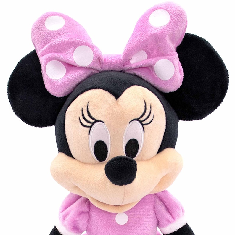 May include: A plush Minnie Mouse doll with a pink bow featuring white polka dots. The doll has black ears, a black nose, and is wearing a pink dress with white trim. The doll's face is beige with black eyelashes and large eyes.