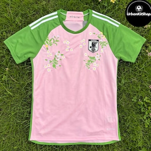 May include: A short-sleeved athletic jersey featuring a light pink center panel with a floral pattern and green side panels with white stripes on the shoulders. The Japan Football Association crest is on the chest, and the text FOGIN is on the collar.