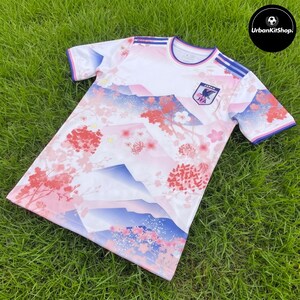 Puede incluir: Una camiseta deportiva de manga corta con base blanca y un estampado degradado de monta&ntilde;as azules y ramas de cerezo en flor rosas. El pecho muestra el escudo de la Asociaci&oacute;n de F&uacute;tbol de Jap&oacute;n con el texto JAPAN y JFA. Rayas azules en los hombros.