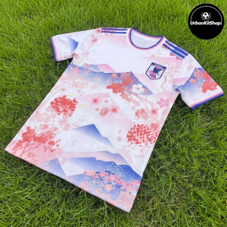 Puede incluir: Una camiseta deportiva de manga corta con base blanca y un estampado degradado de monta&ntilde;as azules y ramas de cerezo en flor rosas. El pecho muestra el escudo de la Asociaci&oacute;n de F&uacute;tbol de Jap&oacute;n con el texto JAPAN y JFA. Rayas azules en los hombros.