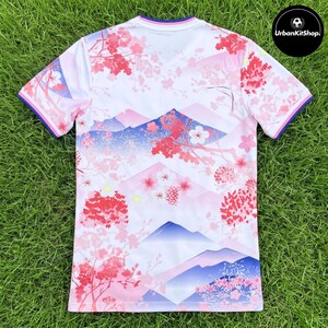 Puede incluir: Una camiseta de manga corta blanca con un patr&oacute;n repetitivo de flores de cerezo rosas y siluetas de monta&ntilde;as azules. El cuello y los pu&ntilde;os de las mangas tienen bandas azules y rosas. El texto UrbanKitShop es visible en un c&iacute;rculo negro.