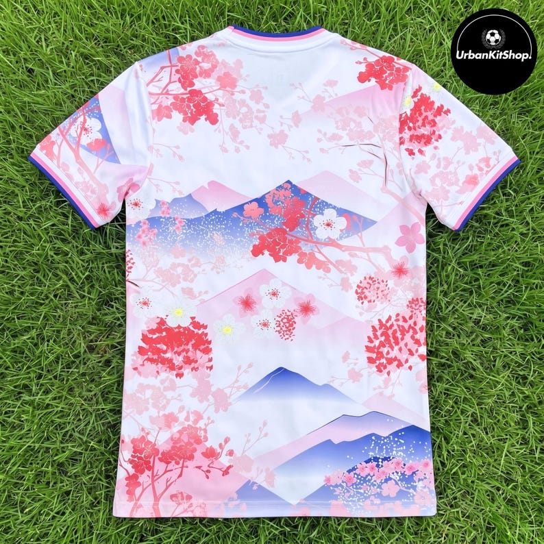 Puede incluir: Una camiseta de manga corta blanca con un patr&oacute;n repetitivo de flores de cerezo rosas y siluetas de monta&ntilde;as azules. El cuello y los pu&ntilde;os de las mangas tienen bandas azules y rosas. El texto UrbanKitShop es visible en un c&iacute;rculo negro.