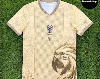 Puede incluir: Una camiseta de cuello en V beige con un panel central punteado, el escudo de la CBF Brasil con cinco estrellas y un gráfico grande de un loro en la parte inferior derecha. Las mangas y los lados tienen motivos florales.