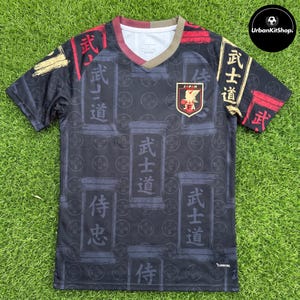 May include: A black short-sleeve jersey featuring a repeating pattern of rectangular panels with Japanese kanji characters. The shirt includes a V-neck collar with gold and red accents, and the Japan Football Association crest on the left chest.