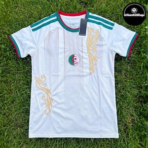 May include: A white short-sleeved soccer jersey featuring green and red trim on the collar and sleeves. The shirt displays a central Algeria emblem, vertical dotted lines, and gold floral patterns on the front. A black tag is attached to the collar.
