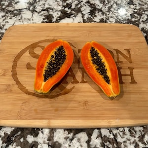 May include: Two halves of a papaya on a wooden cutting board. The board has the words "Saxon Ranch" engraved on it.