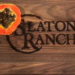 May include: A wooden cutting board with the engraved text "SLATON RANCH" and a half of a papaya. The papaya has a vibrant orange flesh and is filled with black seeds. The cutting board has a rich, brown wood grain.