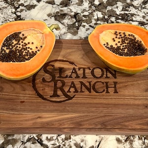 May include: Two halves of a ripe papaya with orange flesh and black seeds rest on a wooden cutting board. The board is engraved with the text "SLATON RANCH" in a rustic font, suggesting a farm or ranch theme.