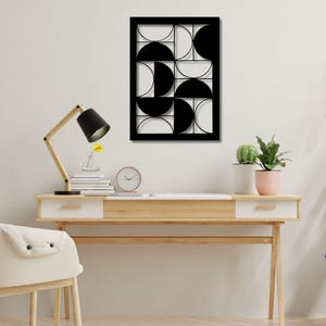 May include: A black and white geometric wall art piece with a rectangular frame. The design features a pattern of semi-circles and quarter-circles. The artwork is mounted on a white wall above a wooden desk with a lamp, clock, and plants.