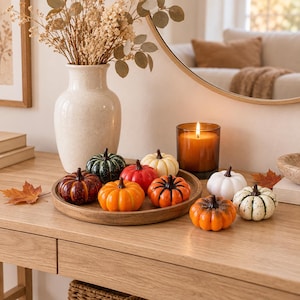Puede incluir: Una colección de pequeñas calabazas decorativas en varios colores, incluyendo naranja, rojo, blanco, verde y patrones moteados. Estas calabazas en miniatura están dispuestas en una bandeja redonda de madera junto a una vela encendida.