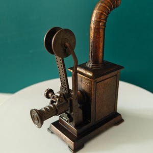 May include: A detailed bronze-colored miniature film projector. The model features a square base, a tall curved chimney, and a reel-to-reel film mechanism. The projector is set against a teal background.