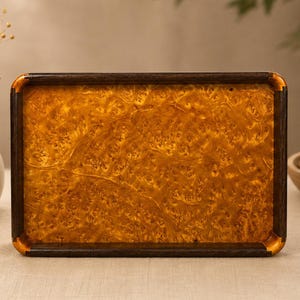 May include: A rectangular serving tray featuring a central panel of golden-brown burl wood with a complex, swirling grain pattern. The tray is framed by a dark, solid wood border with rounded corners, creating a distinct contrast in color and texture.