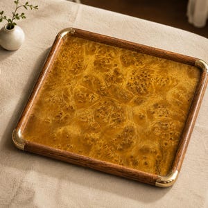 May include: A square serving tray featuring a golden-brown burl wood centre panel with a distinct natural grain pattern. The tray is framed by a dark wood border with polished gold-tone metal corner accents at each of the four rounded corners.