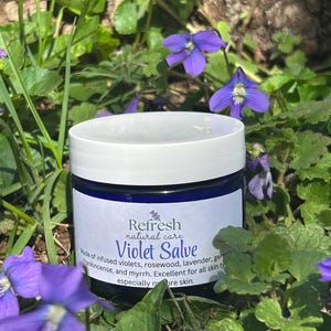 Pode incluir: Um frasco de Violet Salve da Refresh natural care com tampa branca e rótulo azul. O rótulo inclui o texto "Violet Salve" e os ingredientes. O frasco é rodeado por folhagem verde e violetas roxas.