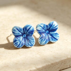 Peut inclure: Une paire de boucles d'oreilles à tige en forme de fleurs à quatre pétales. Chaque boucle présente une finition brillante et émaillée avec un motif aquarelle bleu et blanc, mettant en valeur des nervures de pétales et un centre en relief.