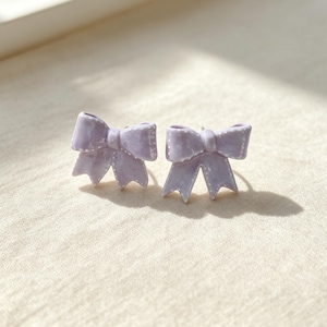 May include: A pair of stud earrings shaped like bows in a light lavender colour. Each bow features a glossy finish with small, white dotted details along the edges, creating a textured appearance on the surface of the lavender-coloured material.