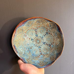Peut inclure: Un bol en céramique rond présentant un motif complexe en relief rappelant un mandala sur toute sa surface. L'émail affiche un mélange de tons turquoise vif, bleu profond et brun cuivré, avec une finition texturée soulignant le design.
