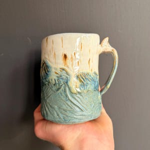Peut inclure: Une tasse en céramique avec une base bleu clair texturée ornée de motifs sculptés en forme de vagues, surmontée d'une section supérieure lisse de couleur crème avec des coulures brunes verticales et une anse incurvée sur le côté droit.