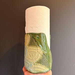 Pode incluir: Um suporte cilíndrico de cerâmica para papel toalha com um esmalte verde brilhante e uma superfície texturizada em relevo. A base apresenta uma mistura de tons verdes claros e escuros com um design geométrico intrincado em relevo.