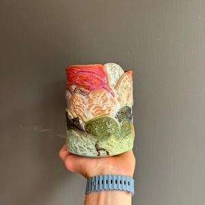 May include: A cylindrical ceramic mug featuring a textured, multi-coloured landscape design. The surface displays layers of red, orange, white, and green glazes with a raised, embossed finish that depicts rolling hills and abstract terrain patterns.