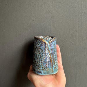 Puede incluir: Un vaso cilíndrico de cerámica con un patrón texturizado similar a escamas de reptil en tonos azules y marrones. El recipiente presenta un diseño distintivo de borde doblado con un esmalte de bronce metálico que acentúa los bordes y textura.