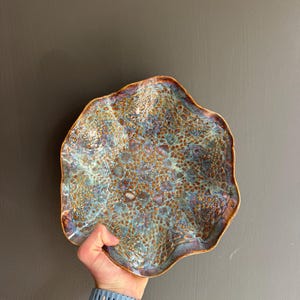 Op de afbeelding: Een ronde keramische kom met een onregelmatige, golvende rand. Het oppervlak heeft een complex, getextureerd patroon in tinten van metallic brons, diepblauw en iriserend paars, wat zorgt voor een gevlekt, organisch uiterlijk over de gehele binnenzijde.