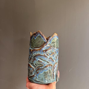 Może przedstawiać: Cylindryczny ceramiczny wazon o teksturowanej, nierównej krawędzi i wykończeniu z reaktywnego szkliwa. Powierzchnia ukazuje mieszankę głębokiej leśnej zieleni, przygaszonego błękitu i ciepłych miedziano-brązowych odcieni, tworząc warstwowy, organiczny wygląd.