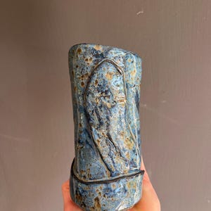 Może przedstawiać: Ręcznie robiony ceramiczny wazon z teksturowanym, niebieskim szkliwem i brązowymi akcentami. Wazon posiada wytłoczony wzór i ciemny druciany detal wokół środka. Wazon jest trzymany w dłoni.