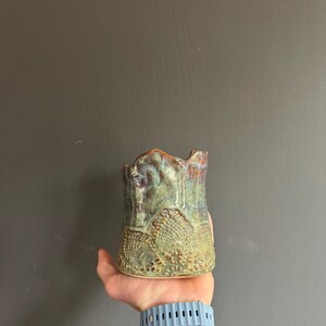 May include: A handmade ceramic vase with a textured, lace-like pattern around the base. The vase has a unique, irregular top edge and is glazed in shades of green, blue, and brown. The vase is held in a hand.