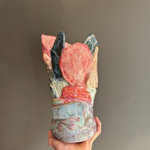 Puede incluir: Un jarrón de cerámica hecho a mano con una forma orgánica e irregular y un esmalte brillante multicolor. La superficie muestra una mezcla vibrante de tonos rosa, rojo, azul y crema con secciones texturizadas y un borde dentado e irregular.