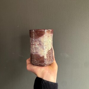Puede incluir: Un vaso de cerámica hecho a mano con un diseño texturizado y acanalado. La taza presenta una base de color burdeos intenso que se desvanece en un tono blanco crema y lavanda. La forma cilíndrica se sostiene en una mano sobre un fondo gris.