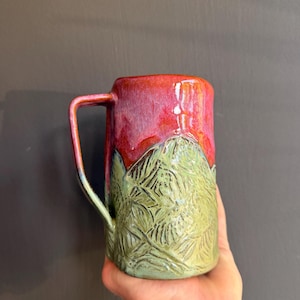 Puede incluir: Una taza de cerámica con una sección superior esmaltada en rojo vibrante y una sección inferior verde texturizada con un patrón de hojas en relieve. La taza tiene un cuerpo cilíndrico alto y un asa angular única en el costado.