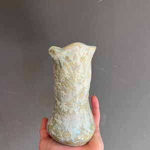 Op de afbeelding: Een handgemaakte keramische vaas met een uniek, getextureerd oppervlak. De vaas heeft een bolvormige basis en een uitlopende, gerimpelde bovenkant. De kleuren zijn gebroken wit, goud en lichtblauw, waardoor een gemarmerd effect ontstaat. De vaas wordt in een hand gehouden tegen een grijze achtergrond.