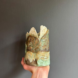 Może przedstawiać: Cylindryczne naczynie ceramiczne z nierówną, poszarpaną górną krawędzią. Powierzchnia pokryta jest nakrapianym szkliwem w odcieniach ziemistego brązu, szałwiowej zieleni i kremu, z teksturowanym, falistym wykończeniem o organicznym wyglądzie.