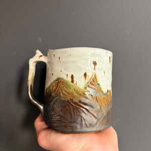 Puede incluir: Una taza de cerámica con un diseño de paisaje montañoso texturizado. La parte superior tiene un esmalte liso de color blanco roto, mientras que la sección inferior muestra tonos marrón oscuro y naranja terroso con líneas talladas.