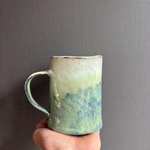 Może przedstawiać: Cylindryczny ceramiczny kubek o teksturowanej, nierównej powierzchni. Górna część pokryta jest gładkim, kremowobiałym szkliwem, podczas gdy dolna sekcja przechodzi w nakrapiane wykończenie w kolorze szałwiowej zieleni i błękitu, z zakrzywionym uchem.