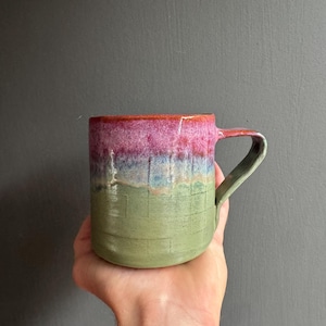 May include: A ceramic mug featuring a multi-coloured glaze with a vibrant pink top section, a light blue middle band, and a sage green base. The surface of the mug displays a subtle, embossed grid pattern throughout, complemented by a matching green handle.