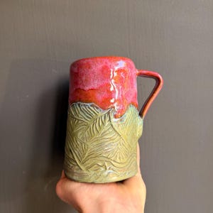 Puede incluir: Una taza de cerámica con una sección superior esmaltada en rosa y rojo vibrante y una sección inferior de color verde salvia texturizada con un patrón de hojas en relieve. La taza tiene un asa en el lado derecho y un cuerpo cilíndrico brillante.