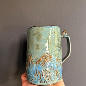 Può includere: Una tazza cilindrica in ceramica con una smaltatura lucida maculata azzurra e verde. La parte inferiore presenta un design a catena montuosa intagliato e testurizzato con accenti marroni. Ha un manico robusto con una piccola sporgenza triangolare.