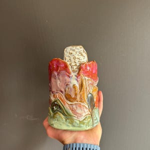 May include: A cylindrical ceramic vessel with a glossy, multi-coloured glaze featuring layers of sage green, burnt orange, deep red, and cream. The rim has an irregular, wavy edge with a central raised, textured, speckled beige tab extending upwards.