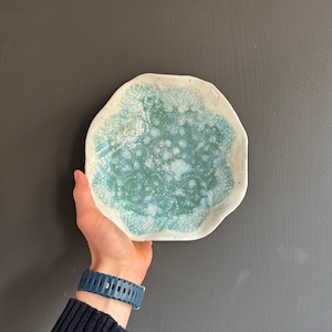 May include: A handmade ceramic plate with an irregular shape. The plate has a turquoise and white glaze, with a speckled pattern in the centre and a white rim. The plate is held against a dark grey background.