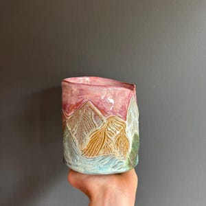 May include: A cylindrical ceramic mug featuring a hand-carved mountain landscape design. The surface displays a gradient of colours including a pink upper section, golden-brown mountain peaks, and a light blue base with textured carved lines.