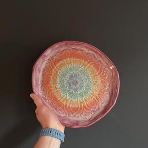 Peut inclure: Une assiette en céramique ronde présentant un glaçage dégradé aux couleurs vives, passant d'un vert pâle central à l'orange et au rose profond sur les bords. La surface présente une texture détaillée en relief rappelant une dentelle.