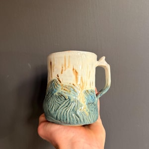 Könnte beinhalten: Ein Keramikbecher mit texturiertem, handgeformtem Körper, der einen cremefarbenen oberen Bereich mit braunen vertikalen Tropfen und einen unteren Bereich mit einem blauen, wellenartigen Reliefmuster aufweist. Der Becher hat einen stabilen Henkel.