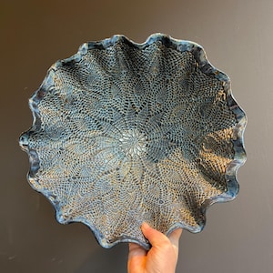 May include: A handcrafted ceramic serving bowl with a unique lace-like pattern in shades of blue, brown, and white. The bowl has a ruffled edge and is held by a hand against a dark background. A decorative piece for the home.