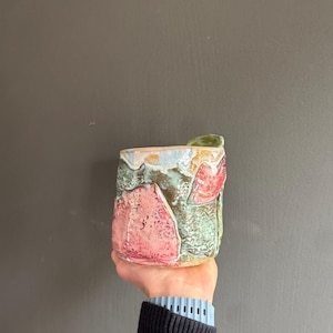 May include: A handcrafted ceramic mug with a textured finish. The mug showcases a mix of pink, green, and blue hues, with an unusual, organic pattern. The mug is held up against a grey backdrop.