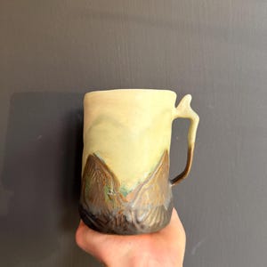 Può includere: Una tazza in ceramica con corpo cilindrico e un manico unico caratterizzato da una piccola sporgenza sulla parte superiore. La tazza presenta una sezione superiore liscia giallo pallido che sfuma in una base testurizzata marrone e verde.