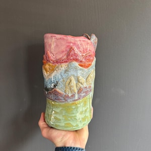 May include: A handcrafted ceramic mug with a landscape design. The mug features layers of pink, orange, blue, and green. The mug has a handle and a textured surface, adding to its unique, artistic appeal. The mug's height is approximately 15 cm.