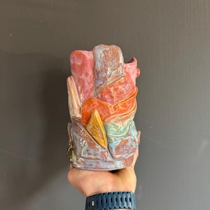 Op de afbeelding: Een kleurrijke keramische vaas met een abstract ontwerp. De vaas heeft een mix van kleuren, waaronder rood, oranje, geel, blauw en paars. Het getextureerde oppervlak voegt visuele interesse toe aan het handgemaakte aardewerk.